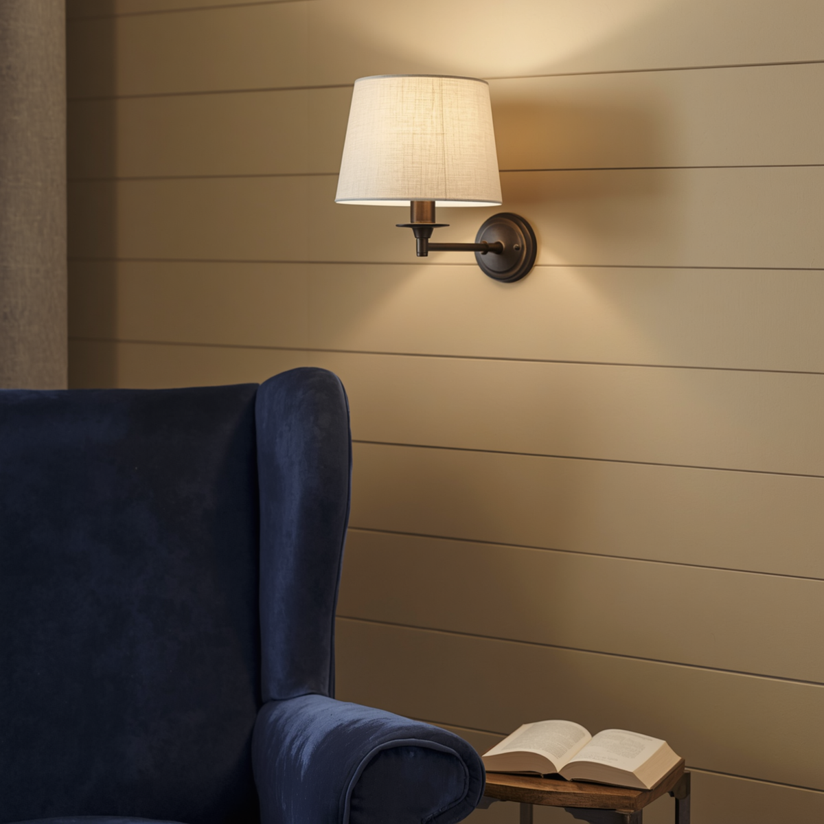 A dark bronze wall sconce with a tapered fabric shade mounted on a shiplap wall above a blue armchair and an open book.