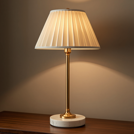 A classic white marble base tall gold table lamp with a pleated fabric shade placed on a dark wooden surface.   